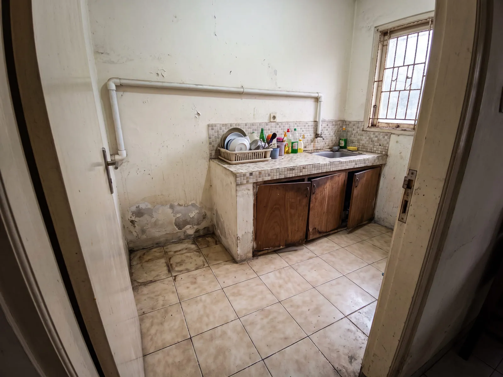 Dapur lama berukuran sempit dengan meja beton berkeramik kecil, pintu kabinet kayu yang melengkung, cat dinding mengelupas di dekat lantai, dan pipa air terpasang di luar dinding.