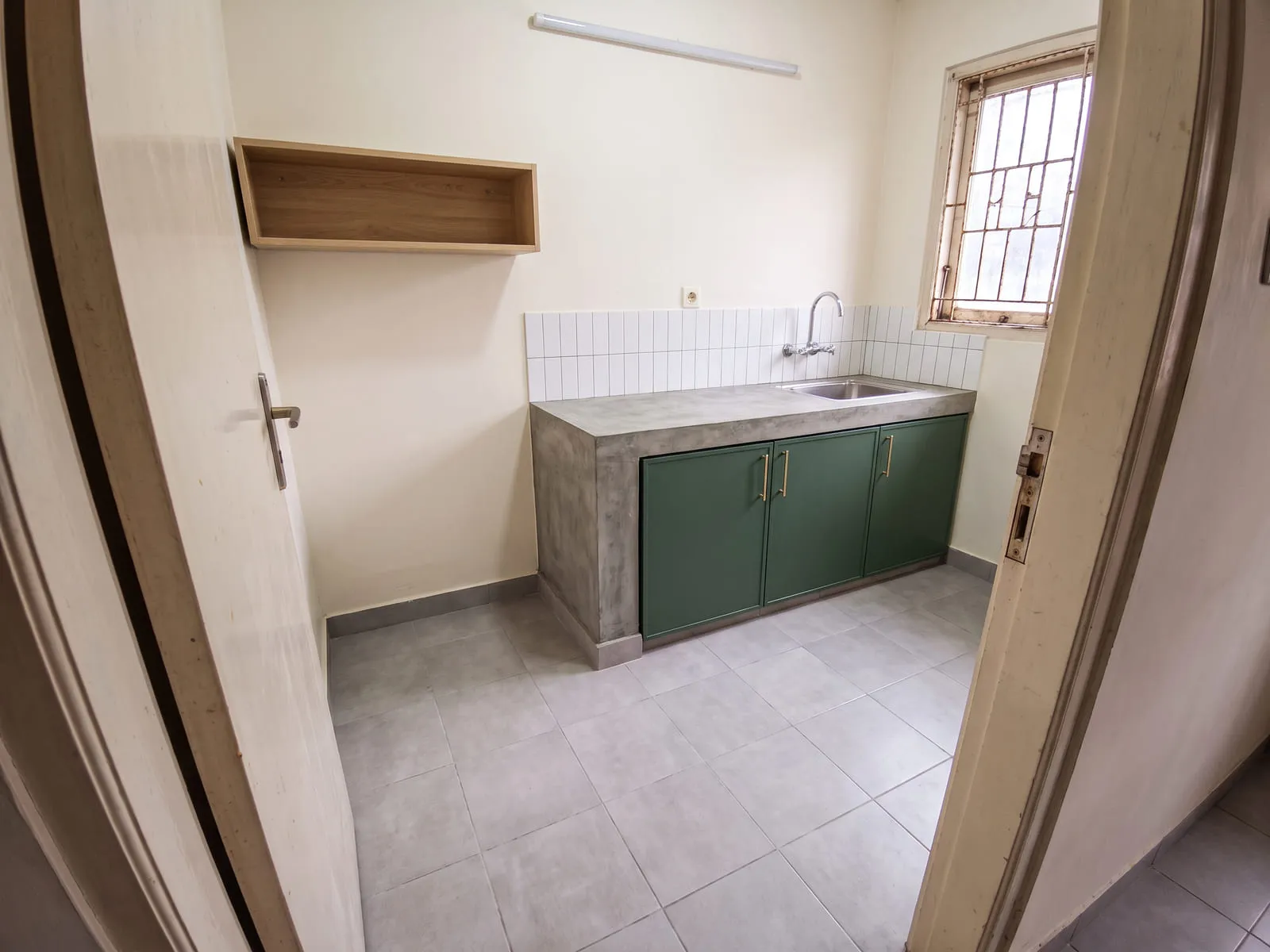 Dapur yang sama setelah dikerjakan, dengan lantai keramik abu-abu, meja beton berpenutup rata, kabinet bawah berwarna hijau tua, backsplash keramik putih, dan pipa yang sudah masuk ke dalam dinding.