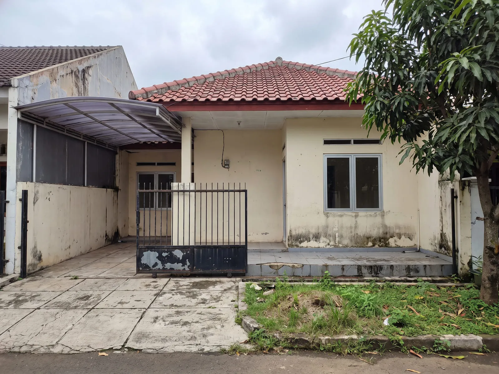 Tampak depan rumah satu lantai dengan cat krem yang kusam dan bernoda di bawah jendela, carport beratap plastik gelombang, teras berkeramik lama, dan pagar besi yang catnya mengelupas.
