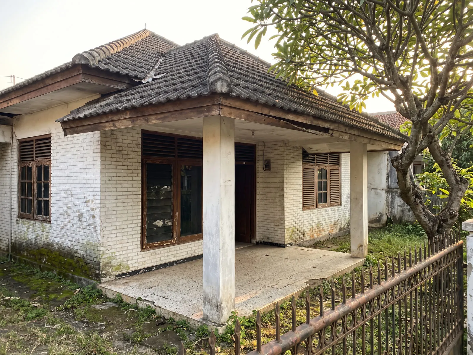 Tampak depan rumah 1980-an dengan dinding bata dicat putih yang sudah berlumut di bagian bawah, jendela kayu krepyak yang pernisnya mengelupas, teras berlantai teraso retak, dan lisplang atap yang melengkung.