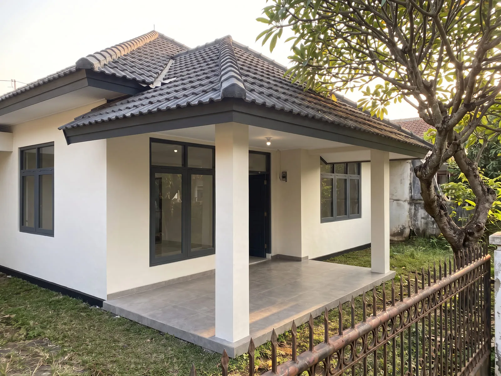 Rumah yang sama setelah dikerjakan, dengan dinding diplester ulang dan dicat putih hangat, jendela berbingkai abu-abu tua di bukaan yang sama, teras berkeramik besar, dan atap bergenteng serta lisplang baru.