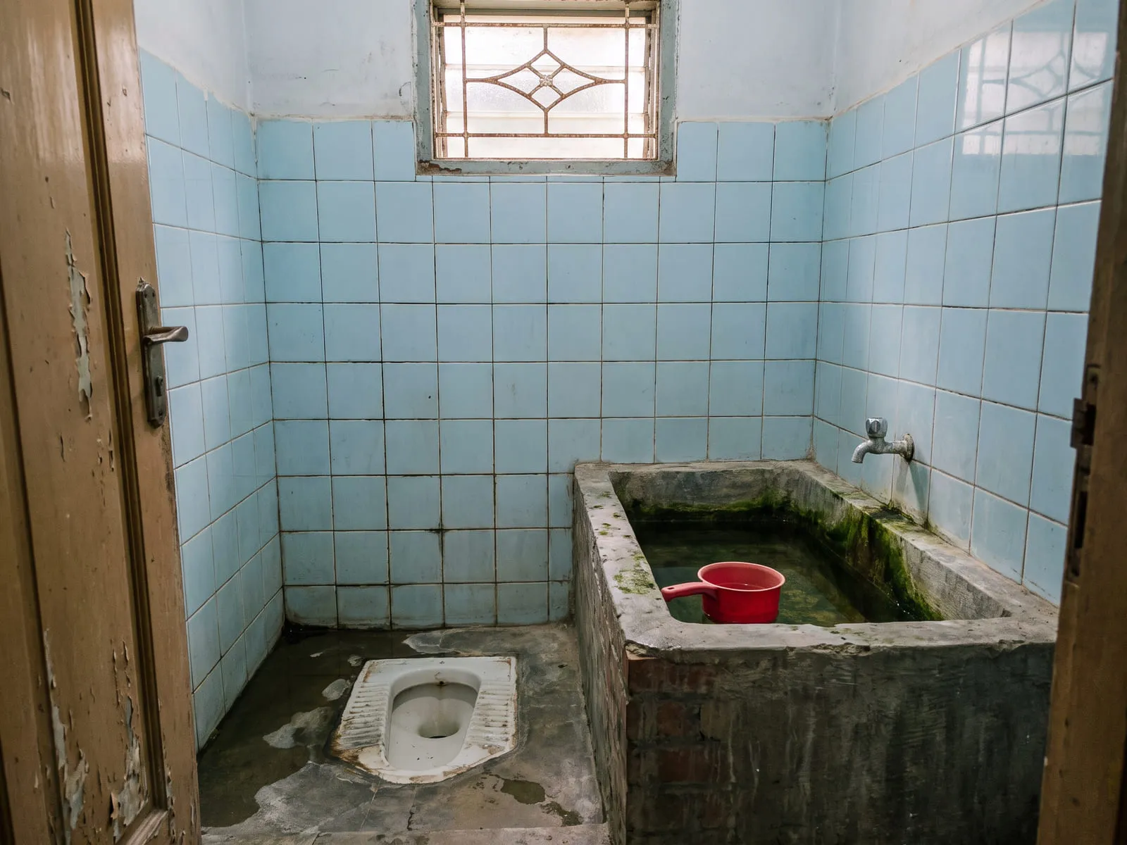 Kamar mandi lama dengan keramik dinding kecil bernat menghitam, kloset jongkok, dan bak air pasangan bata yang sudah berlumut di bagian dalam.