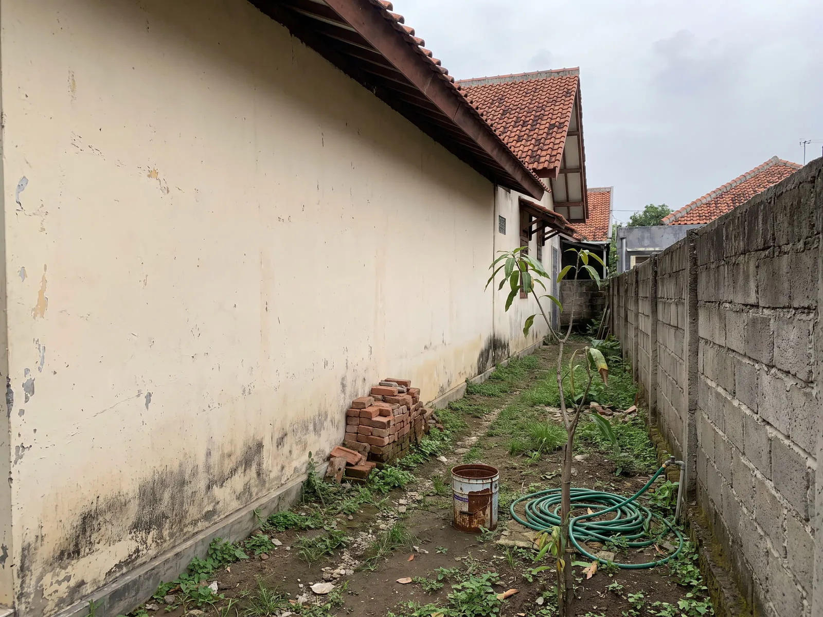 Sisi samping rumah dengan tanah kosong, dinding samping tanpa jendela, dan sisa material bangunan lama di dekat pagar.