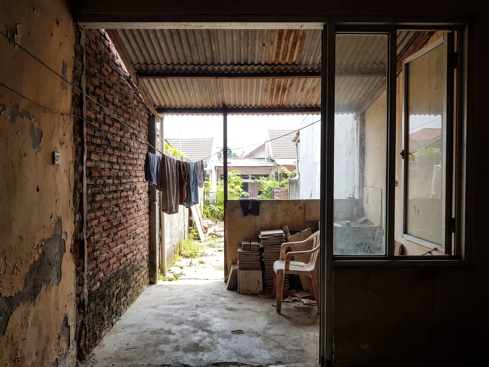 Area servis belakang rumah yang belum jadi, dengan lantai semen, dinding bata terbuka, atap seng bergelombang, jemuran, dan tumpukan keramik sisa di sudut.